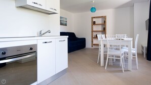 A modern kitchen with white cabinetry, a built-in oven, and a sink. There is a dining area with white chairs and a table, and a blue sofa in the background. - Villa Tola - Blu - One Bedroom Apartment, Sleeps 5 (Valdisotto)