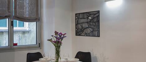 Ein Essbereich mit einem runden Tisch, gedeckt für vier Personen, einer Vase mit violetten Blumen und einem schwarzen, abstrakten Kunstwerk an der Wand.