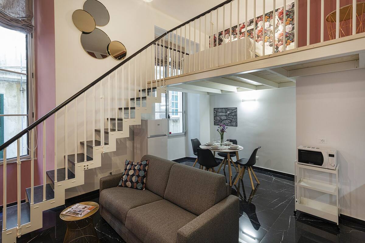 Ein modernes Wohnzimmer mit einem grauen Sofa, einem kleinen runden Tisch und einer Treppe, die zu einer oberen Etage führt.