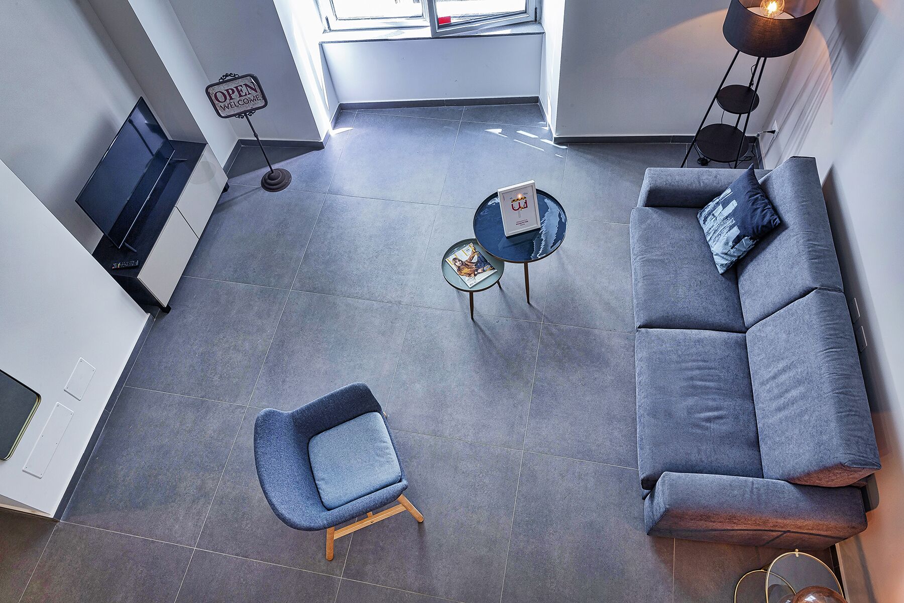 Una sala moderna con un sofá gris, un sillón azul y una mesa de centro con una revista. Se ve un cartel de 'Welcome' y una lámpara de pie.