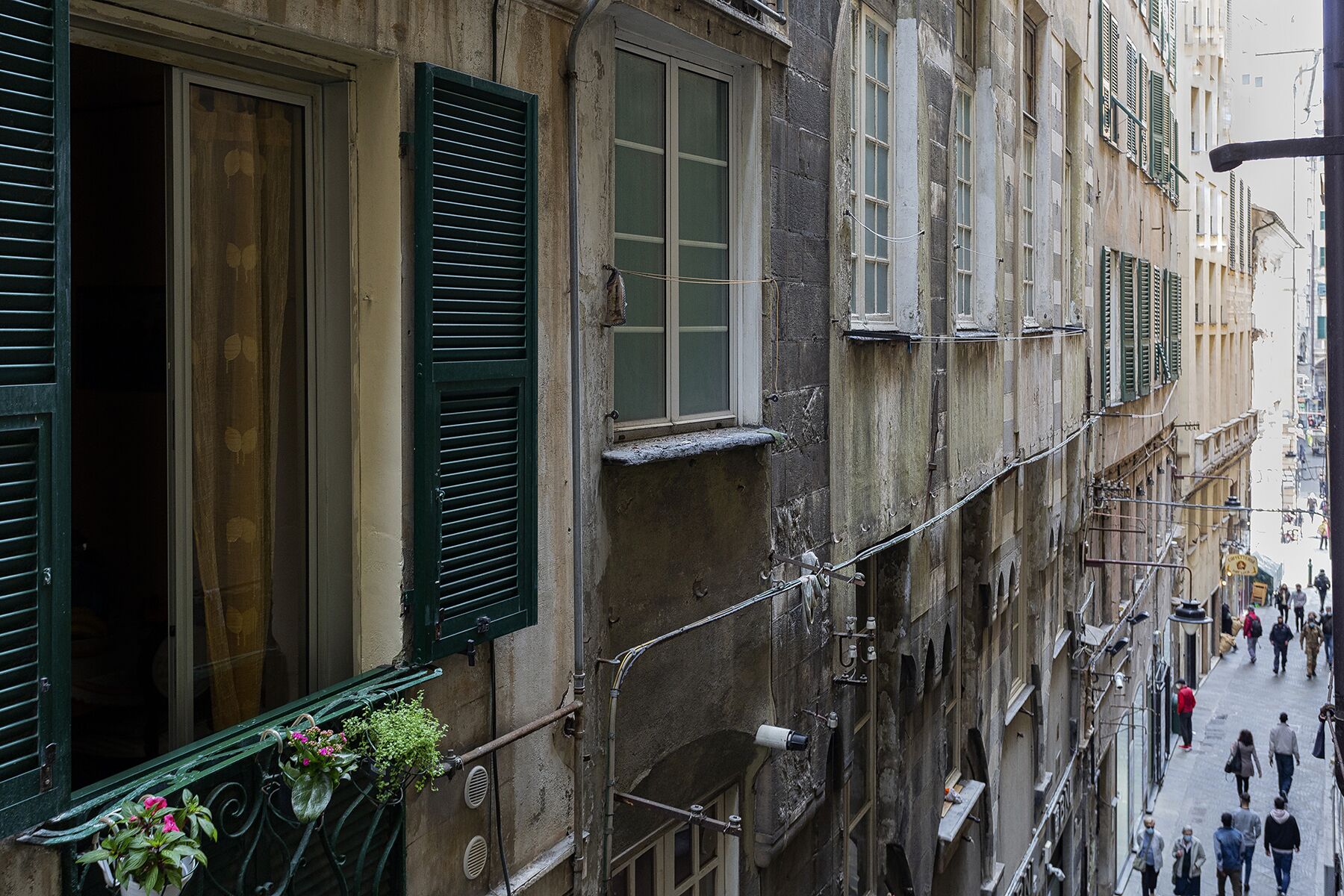 A narrow street with old buildings, some with green shutters, and a few balconies with plants.