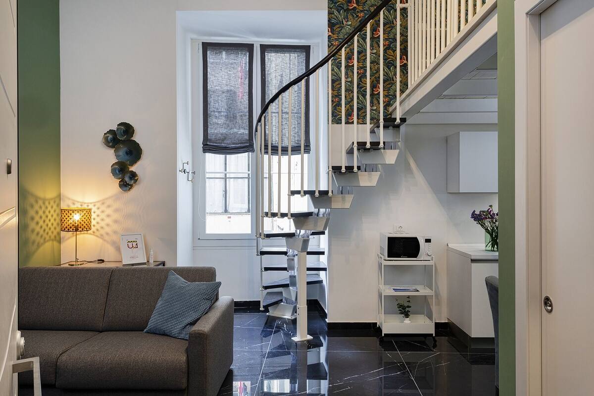 Ein modernes Wohnzimmer mit einem grauen Sofa, einem kleinen Tisch mit Lampe und einer Treppe mit weißer Handläufer, die in einen Küchenbereich führt.