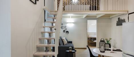 Un apartamento moderno con una escalera que lleva a un dormitorio tipo loft, una zona de comedor con una mesa redonda y una cocina con refrigerador.