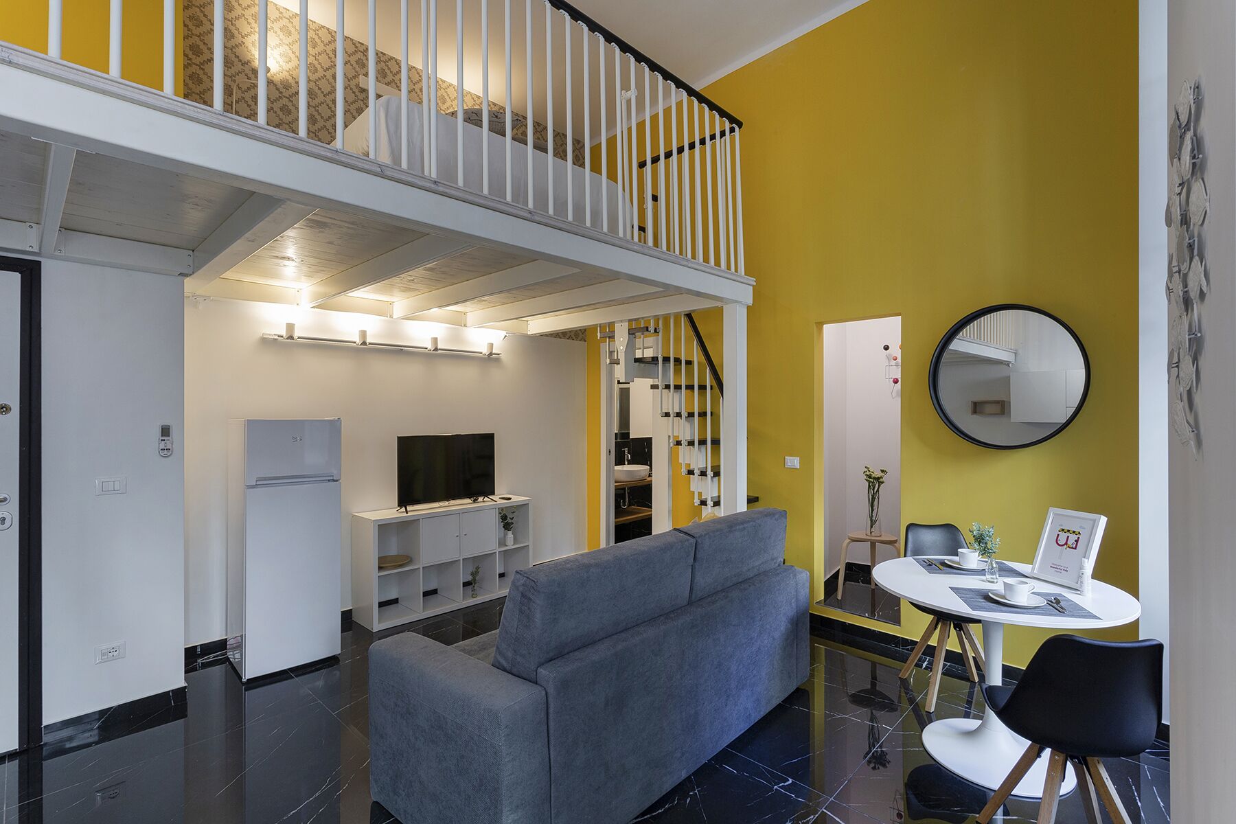 A modern living space with a grey sofa, a dining table with chairs, a TV, and a staircase leading to an upper level.