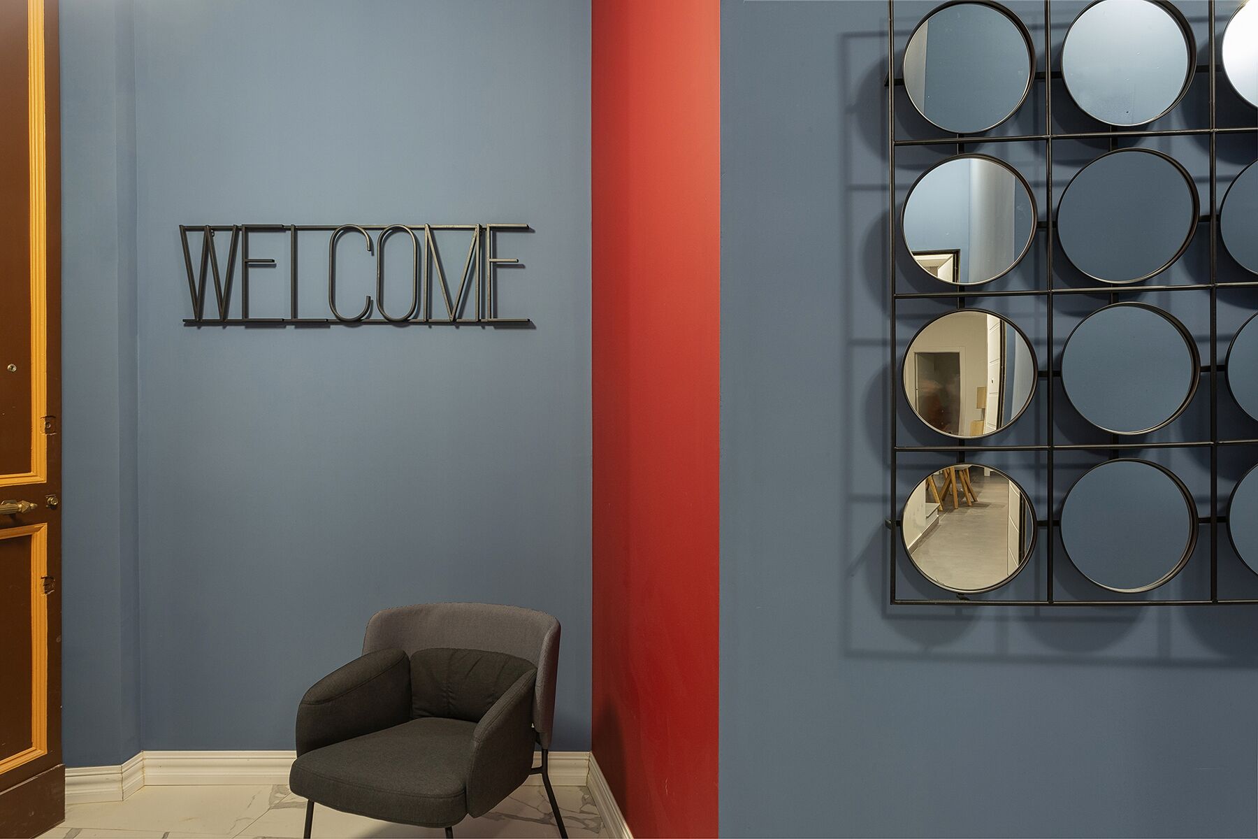 A room with a 'WELCOME' sign, a grey armchair, and a wall with circular mirrors.