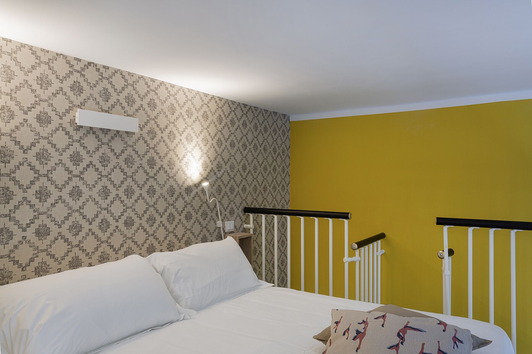 A bedroom with a bed, a nightstand, a wall-mounted lamp, and a yellow accent wall.