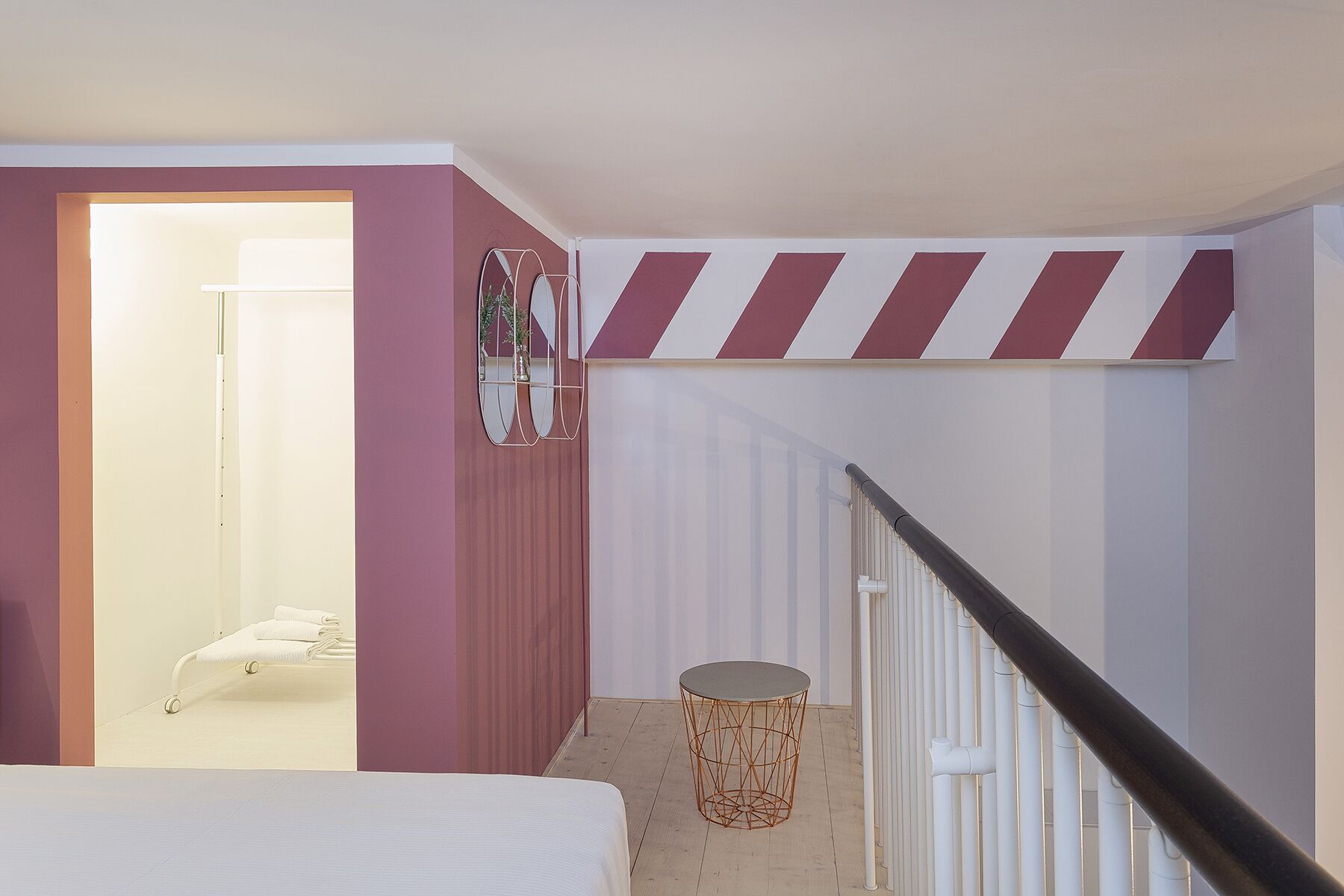 A modern bedroom with a bed, a bedside table, and a mirror. There is a staircase with a striped railing leading to another room.