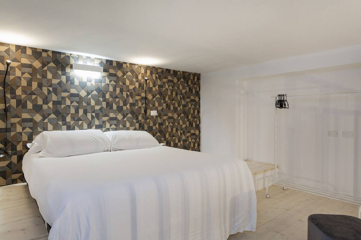 Ein Schlafzimmer mit einem Bett, einer Wand mit geometrischem Muster, einer weißen Tür und einem kleinen Hocker.