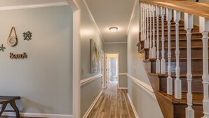 Un pasillo con suelo de madera, una escalera con barandillas blancas y una puerta que da a otra habitación.