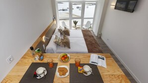 Une chambre d’hôtel moderne avec un balcon, un lit avec des oreillers, une table de petit-déjeuner avec une assiette de fruits, une tasse et un livre.