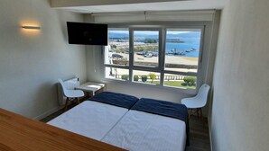 Une chambre à coucher comprenant un lit, un bureau, deux chaises, un téléviseur et une grande fenêtre donnant sur des bateaux et un port.
