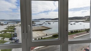 Une vue à travers une fenêtre sur une marina, avec des bateaux, une zone sablonneuse et des collines au loin.