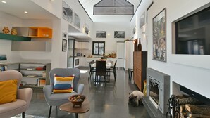 Amplia sala de estar con techo alto, chimenea y comedor con cocina al fondo.
