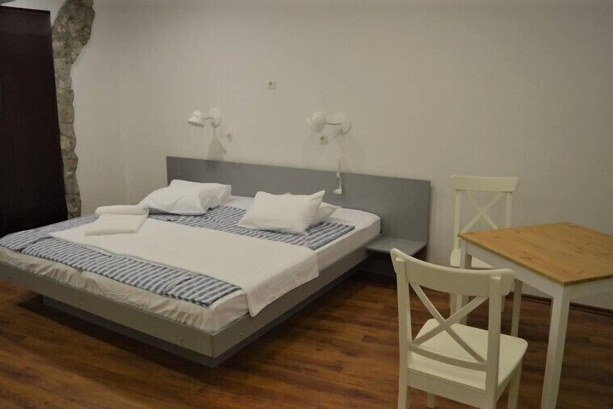 A neatly made bed with white linens, a wooden nightstand, a white chair, and a small wooden table.