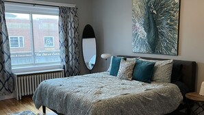 A bedroom with a large bed, a window with curtains, a radiator, a floor lamp, and a peacock painting on the wall. - Large 2 Bedroom 1 Bath Apartment right on Main Street in Downtown Ellsworth. (Ellsworth)