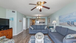 A living room with a ceiling fan, a television, a sofa, a coffee table, and a kitchen in the background. - North Shore Villas 903-3BR/3BA Oceanfront-Ocean Drive (North Myrtle Beach)