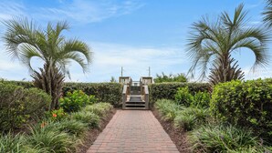 A paved pathway leading to a wooden gate in a landscaped garden with palm trees and dense foliage. - North Shore Villas 903-3BR/3BA Oceanfront-Ocean Drive (North Myrtle Beach)