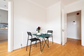 Un coin repas avec une table dressée pour deux personnes, deux chaises, un vase avec des fleurs et, au fond, une cuisine.