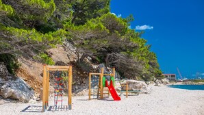 Une aire de jeux de plage avec des structures d’escalade et un toboggan, entourée d’arbres et surplombée d’un ciel bleu clair.