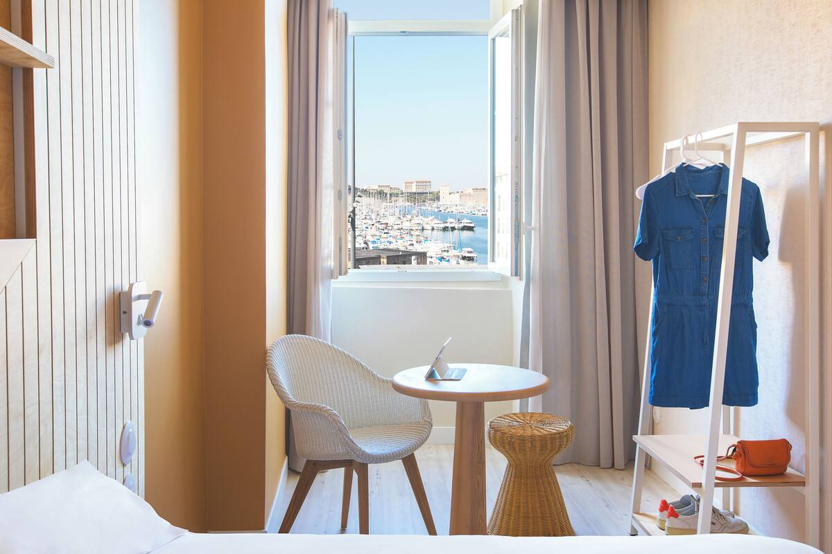 A hotel room with a bed, a chair, a small table with a laptop, a view of a marina, and a clothing rack with a blue dress.