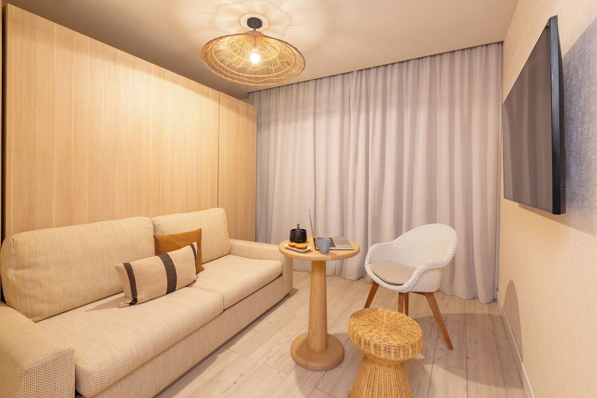 A modern living room with a sofa, a round wooden table, a wicker chair, a woven stool, and a ceiling light fixture.