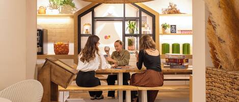 A group of people seated at a wooden table in a cozy, well-lit room with a large woven ceiling light, a window with a view, and shelves with various items.