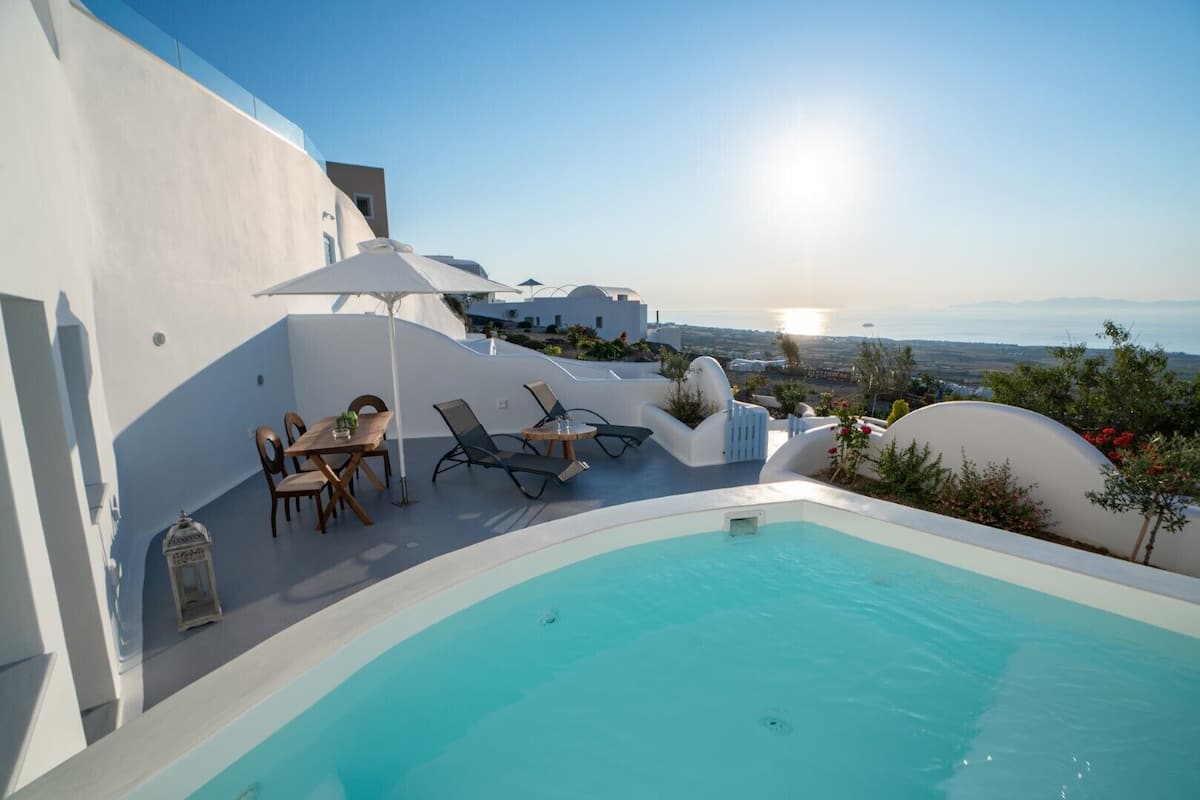 A poolside area with a table and chairs, a sun umbrella, and a view of the sea and landscape.