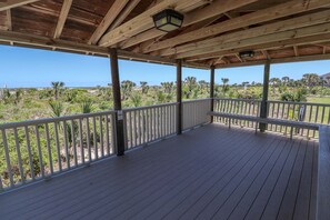 A wooden deck with a railing, overlooking a landscape with palm trees and a distant view of the ocean.
