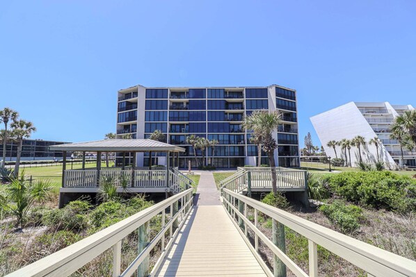 A modern building with a central atrium, a wooden boardwalk leading to it, and a clear sky.