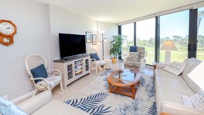 Een woonkamer met een flatscreen televisie, een bank, een salontafel en een groot raam met uitzicht op een grasveld.