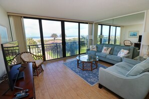 Ein geräumiges Wohnzimmer mit Balkon, einer Couch, einem Couchtisch und einem Schreibtisch mit Computer und Fernbedienungen.