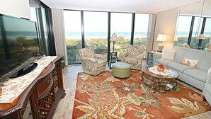 A spacious living room with a large TV, floral armchairs, a sofa, and a view of the ocean. - Anastasia Condos 409- Luxury Direct Oceanfront Fourth Floor Condo (St Augustine)