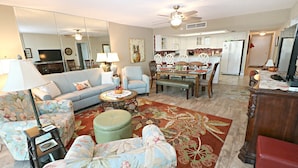 A living room with a sofa, armchair, ottoman, and coffee table. There is a dining area with a table and chairs. The kitchen is visible in the background with cabinets and appliances. - Anastasia Condos 409- Luxury Direct Oceanfront Fourth Floor Condo (St Augustine)