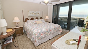 A bedroom with a large bed, two bedside lamps, a nightstand, a wooden headboard, a floral bedspread, a view of the ocean through a large window, and a ceiling fan. - Anastasia Condos 409- Luxury Direct Oceanfront Fourth Floor Condo (St Augustine)