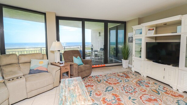 A living room with a sofa, a recliner, a coffee table, a TV, and a balcony with a view of the ocean.