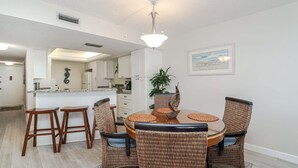 Een eetruimte met een ronde tafel, vier rieten stoelen, een plant in het midden en een keuken op de achtergrond.