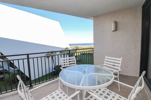 Un balcon avec une table en verre et des chaises blanches, d’où l’on peut admirer un immeuble et une vue sur l’océan.