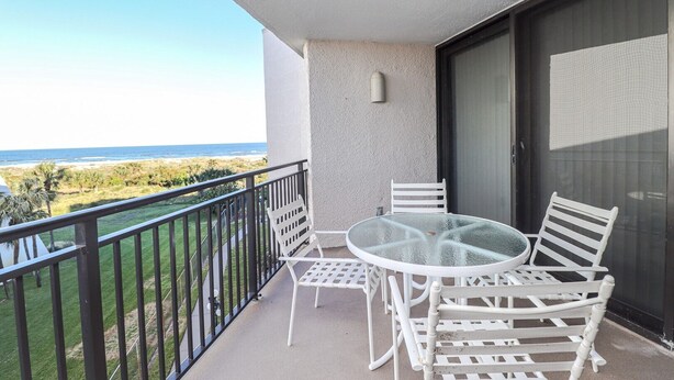 Een balkon met een glazen tafel en witte stoelen, met uitzicht op een strand en de oceaan.