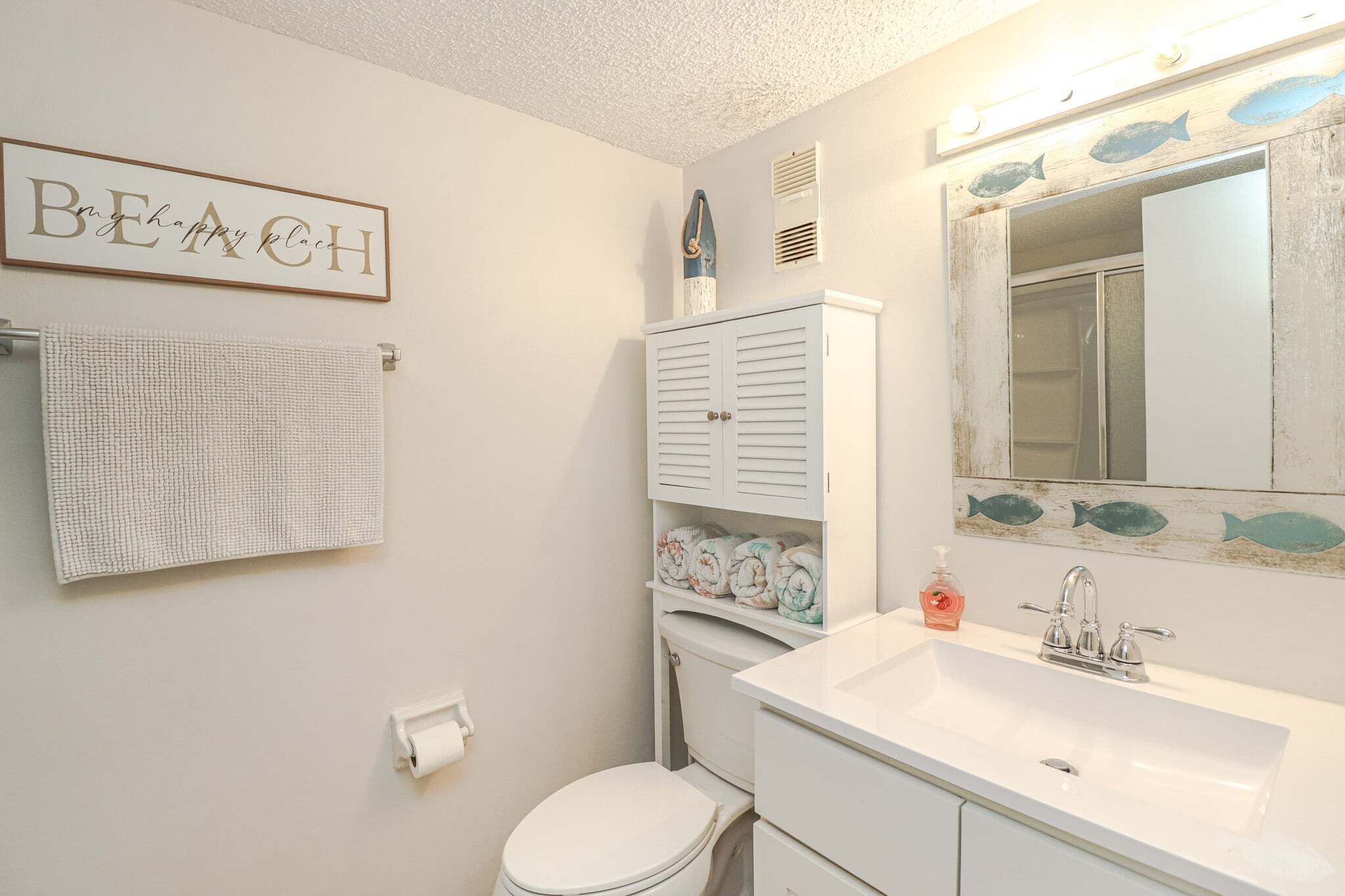A bathroom with a toilet, sink, and cabinets. There is a towel hanging on the wall and a sign with the word 'BEACH' on it.