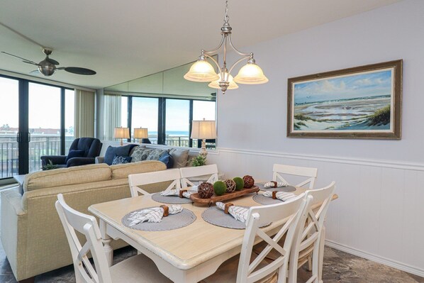 Un coin repas avec une table en bois, des chaises blanches et un lustre. On y trouve également un canapé avec des coussins et un tableau représentant une scène de plage accroché au mur.
