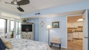 Een slaapkamer met een bed, een televisie, een klok, een lamp en een stoel. Een balkon en een keuken zijn zichtbaar door een geopende deur.