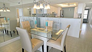 A modern kitchen with a central island, white cabinetry, and a dining area with glass tables and chairs. - Miles of Wide Sandy Beaches! Anastasia Condos 603 (St Augustine)