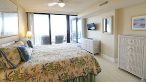 A bedroom with a large bed, a ceiling fan, a television, and a wicker side table. - Miles of Wide Sandy Beaches! Anastasia Condos 603 (St Augustine)