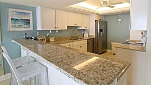 A kitchen with white cabinets, granite countertops, a stainless steel refrigerator, and a ceiling fan. - Miles of Wide Sandy Beaches! Anastasia Condos 603 (St Augustine)