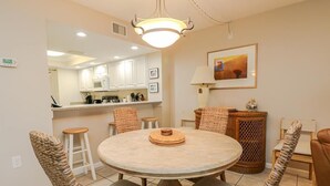 A dining area with a round table, wicker chairs, a lamp, and a kitchen in the background. - Oceanfront Charm in Barefoot Trace 114 (St. Augustine)