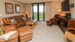 A living room with a brown leather sofa, a brown leather armchair, a wooden coffee table, and a television on a wooden cabinet. There are two framed pictures on the wall, a lamp on the sofa, and a view of a green landscape through the glass door. - Oceanfront Charm in Barefoot Trace 114 (St. Augustine)