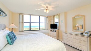 Ein Schlafzimmer mit einem großen Bett, einem Deckenventilator, einem Fernseher, einem Schminktisch und einem großen Fenster mit Blick auf den Strand.