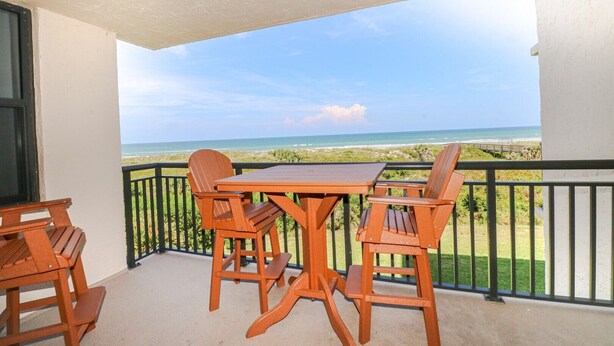 Ein Balkon mit Holztisch und -stühlen, der Blick auf Strand und Meer gewährt.
