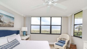 Una camera da letto con un letto, una sedia, un comodino, una ventola a soffitto e un'ampia finestra vista spiaggia.