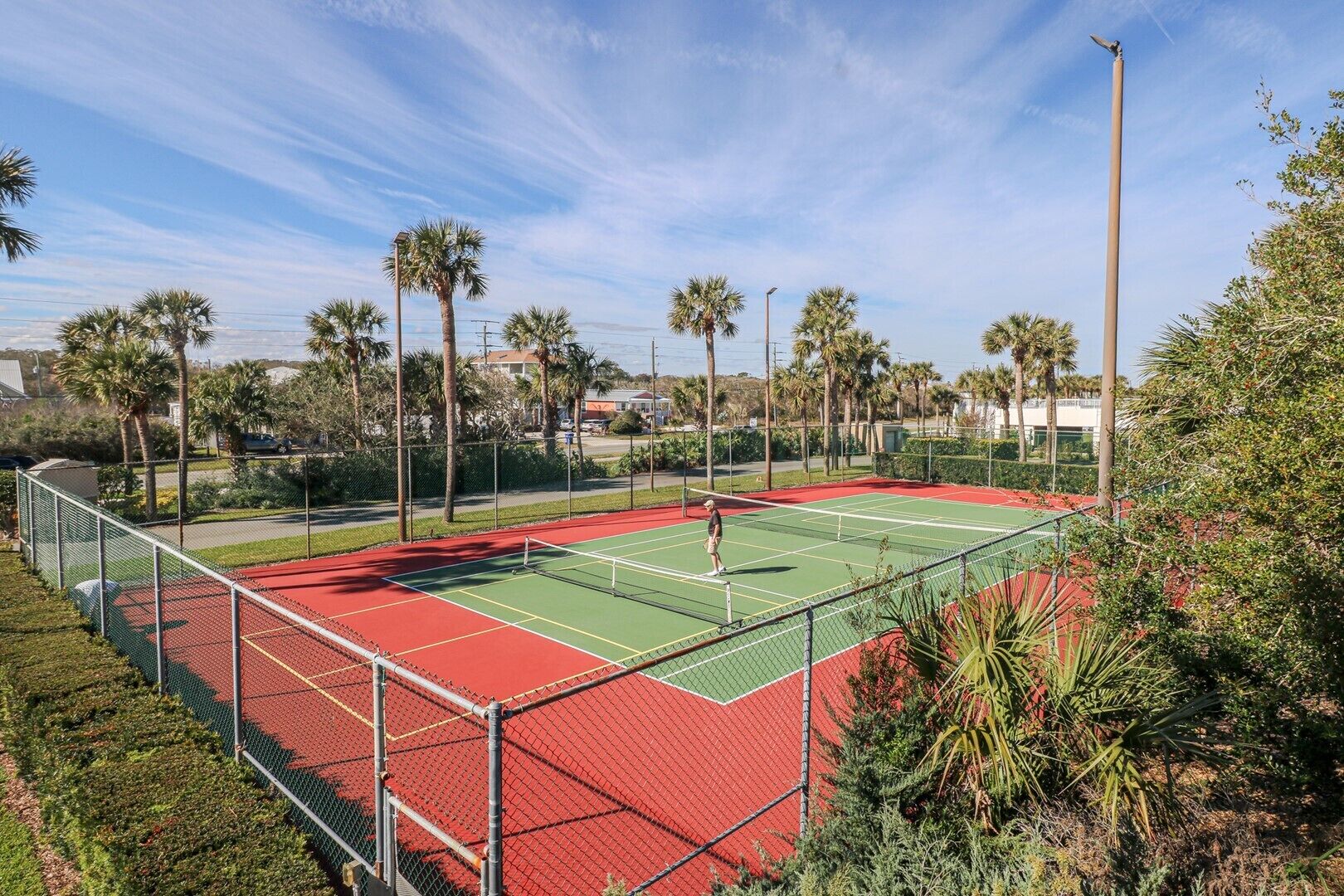 A tennis court with a red and green surface, surrounded by palm trees and a fence.